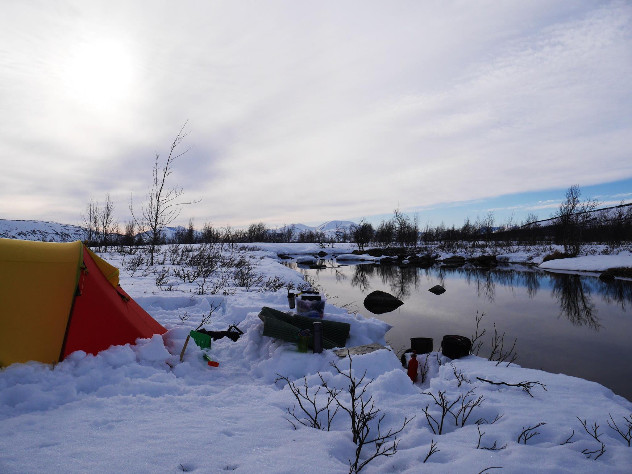 Winter camping scene with a red tent by a snowy river under a cloudy sky.