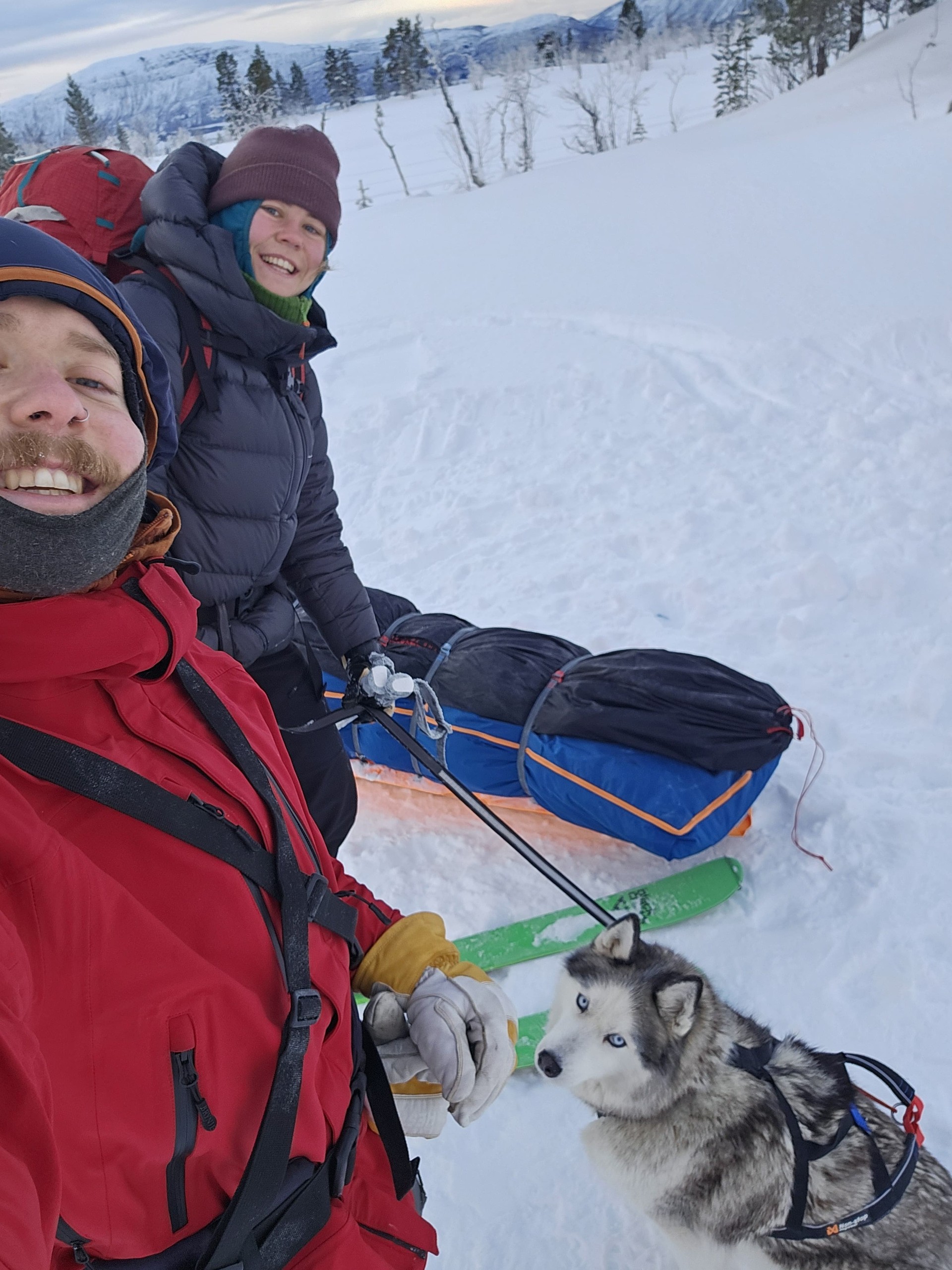 Two people in winter gear with a sled and husky in a snowy landscape.