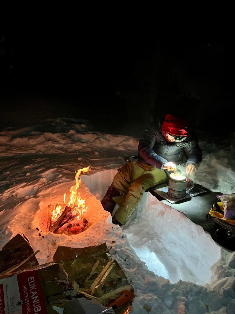 Person in winter gear cooking by campfire in snowy pit at night.