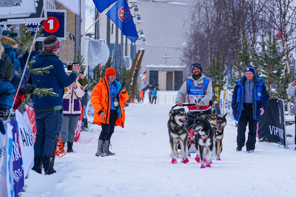 Musher with sled dogs crossing snowy finish line, spectators cheering.