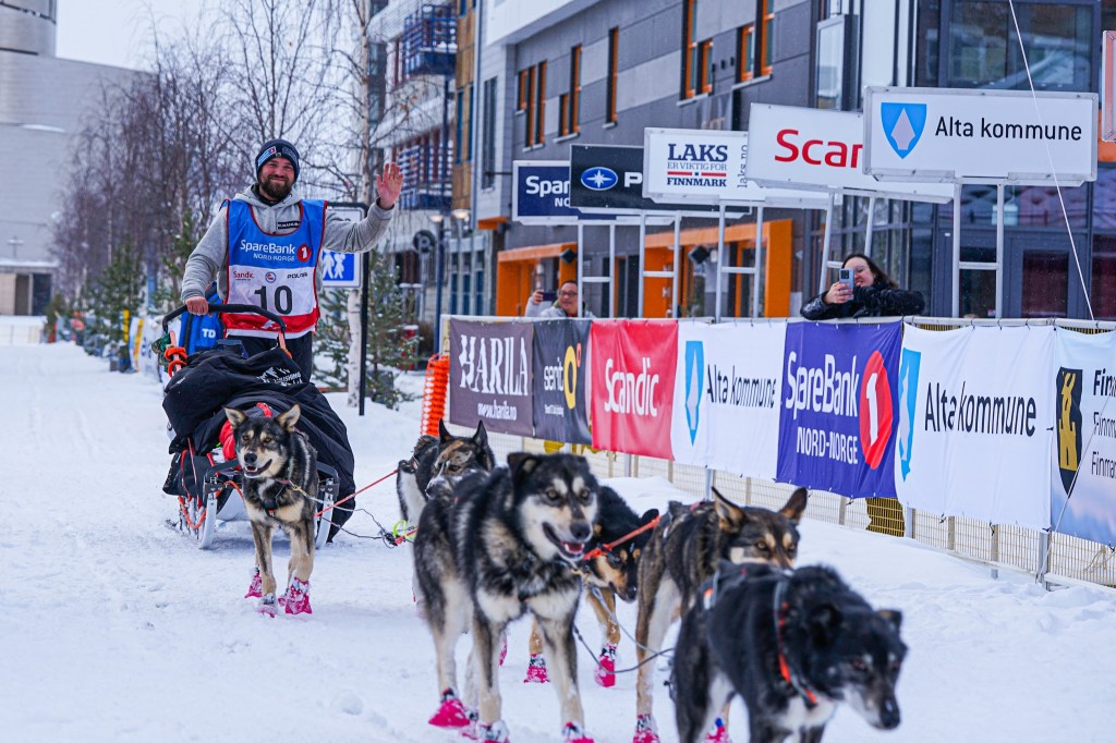 Person on sled with six sled dogs in snow, waving to spectators.