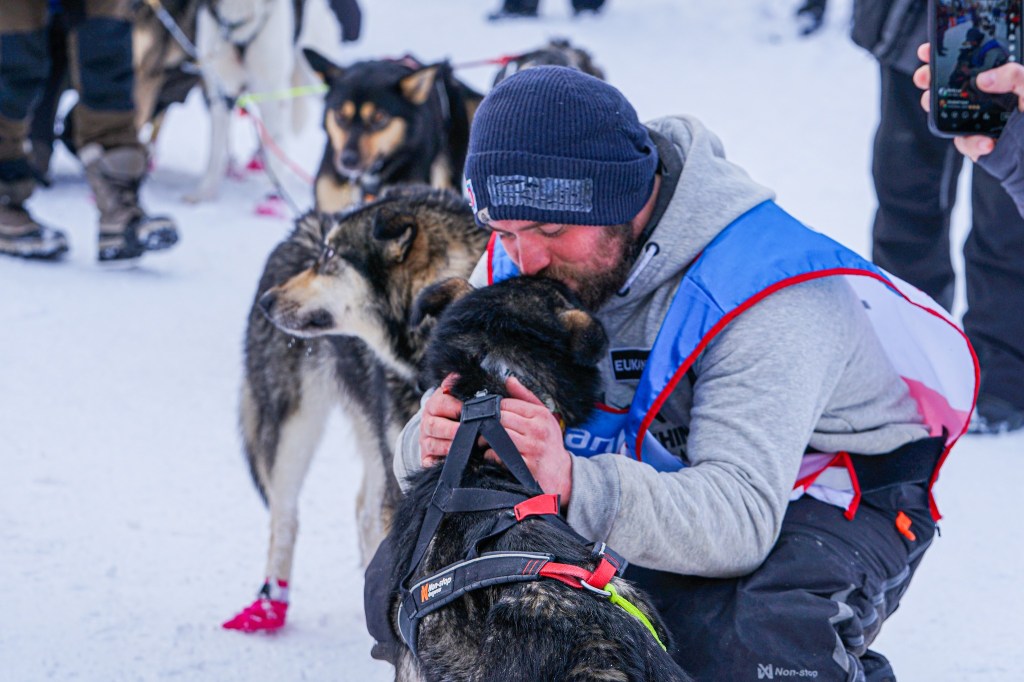 Man in winter gear affectionately hugging a sled dog on snow-covered ground.