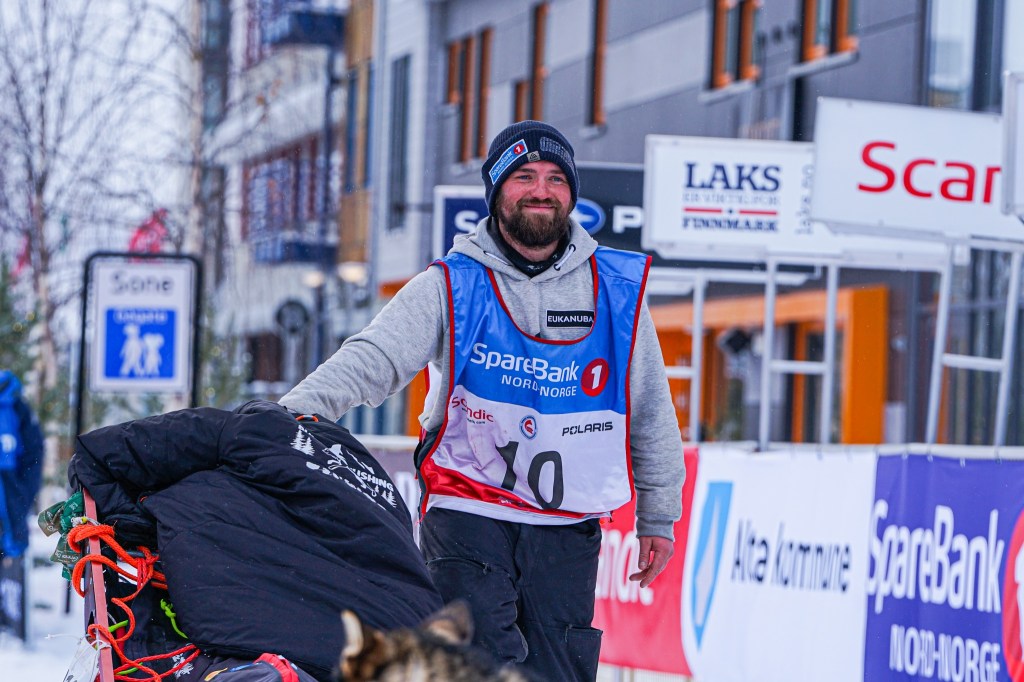 Person smiling with sled and dog, wearing winter gear in snowy, urban setting.