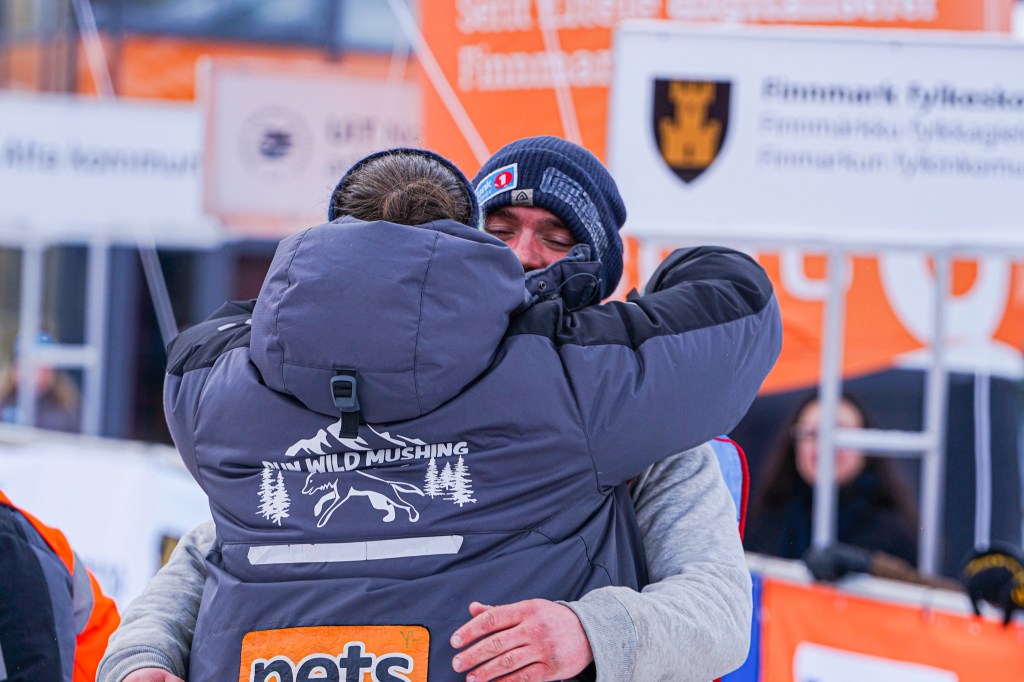 Two people in warm clothing embrace at an outdoor winter event with banners in the background.