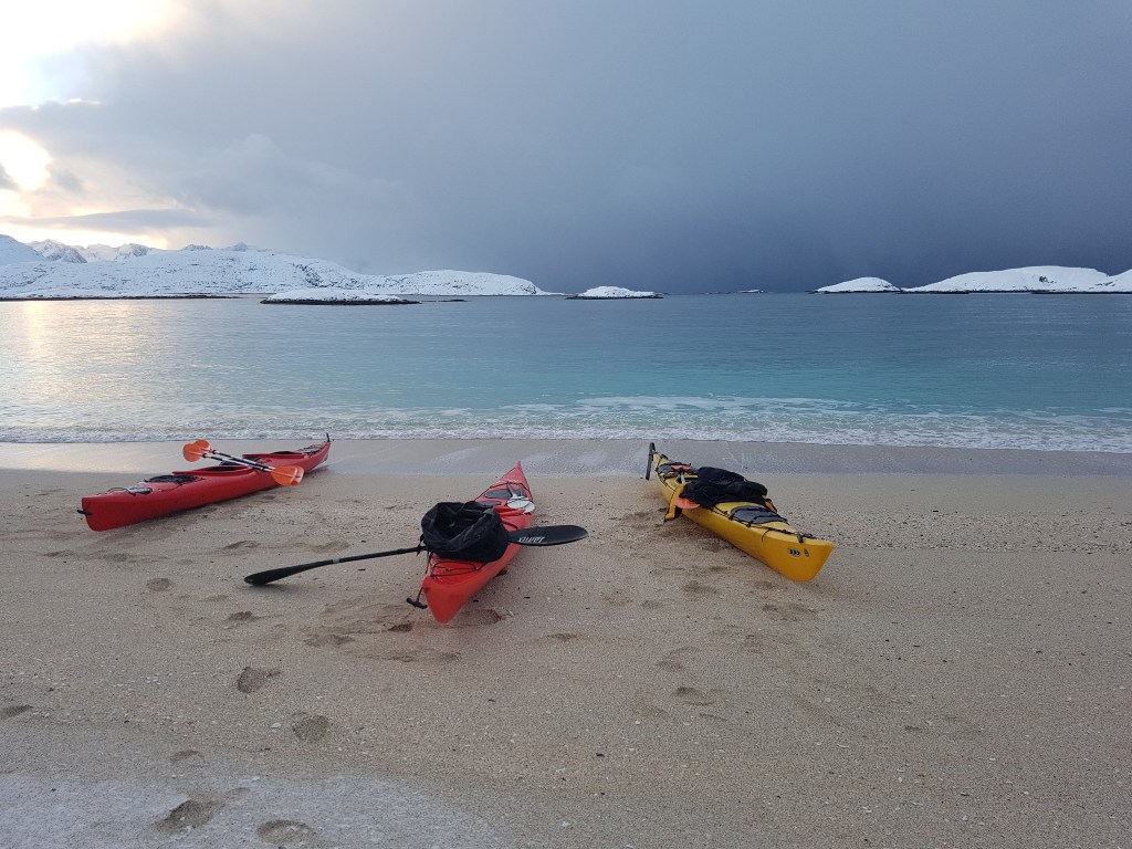 Three kayaks on a snowy beach by calm water under a cloudy sky.