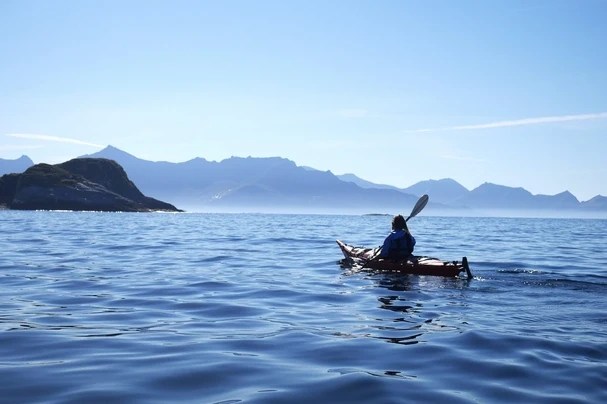 Person kayaking on calm sea with distant mountains under clear blue sky.