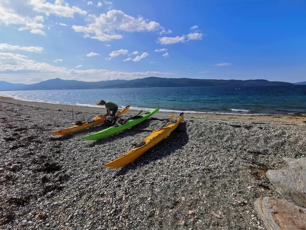 Kayaks on a rocky beach with person kneeling, sea and mountains in the background.