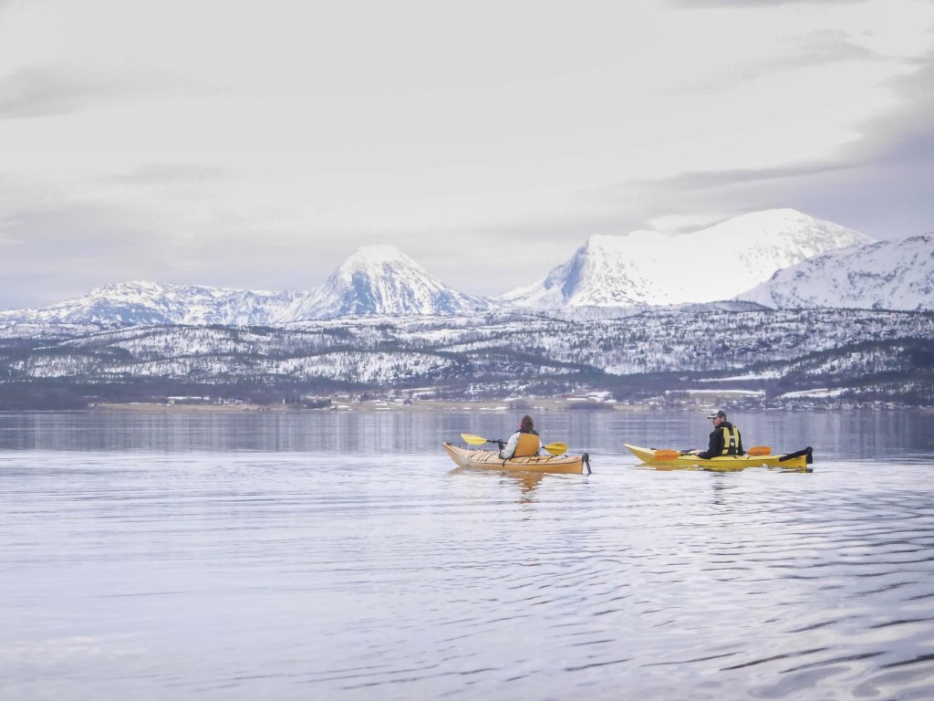 Two kayakers on a calm lake with snow-covered mountains in the background.