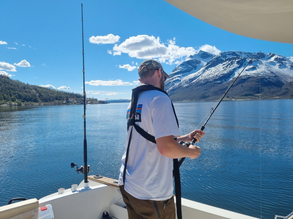 Person fishing on a boat in a lake surrounded by mountains under a clear blue sky.