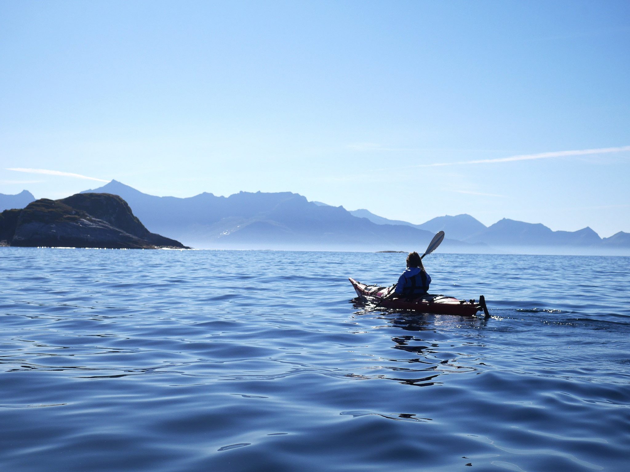 Person kayaking on calm sea with distant mountains and clear sky.
