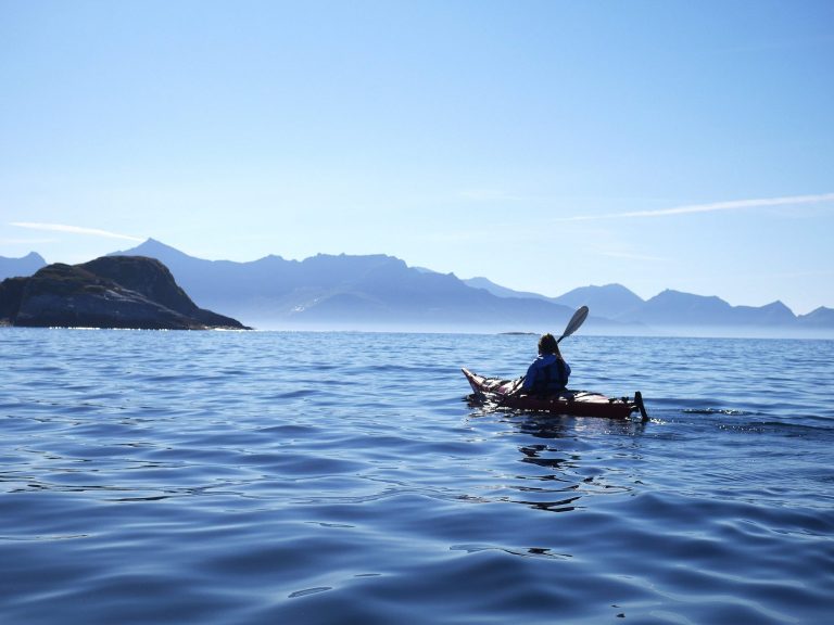 Person kayaking on calm sea with distant mountains and clear sky.