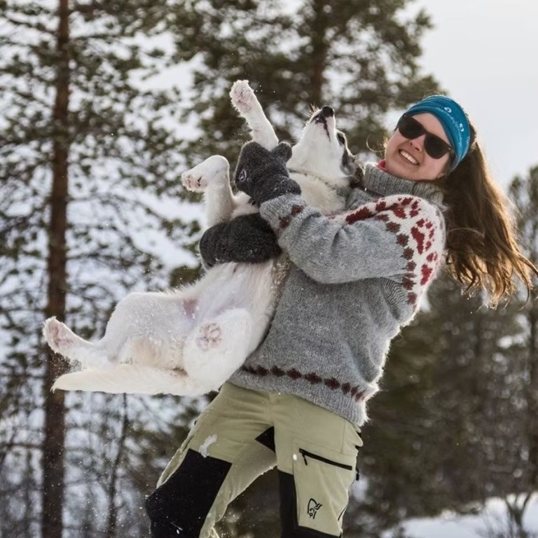 Person in snow holding a playful dog wearing a winter sweater and hat.