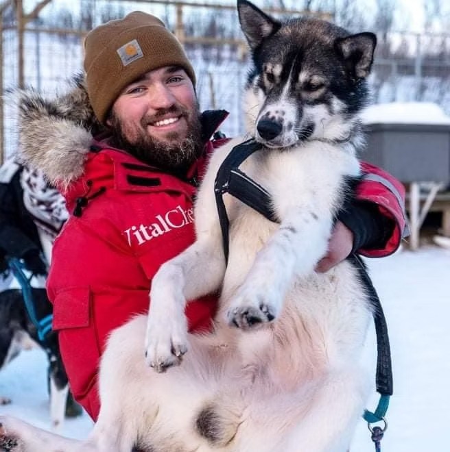 Man in red jacket holds a husky dog outdoors in snowy area.